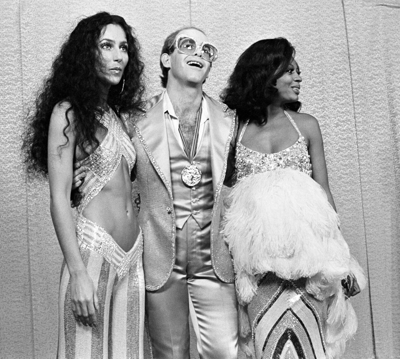 Zwartwit-foto met links zangeres Cher, in het midden zanger Elton John en rechts zangeres Diana Ross. Cher heeft lang zwart haar en draagt een pak met glitters en een driehoekige uitsnede in het midden. Elton John draagt een grote bril en een glimmend pak met een medaille om zijn nek. Diana Ross draagt een glitterjurk met strepen aan de onderkant en in haar hand waarschijnlijk een tas van veren.