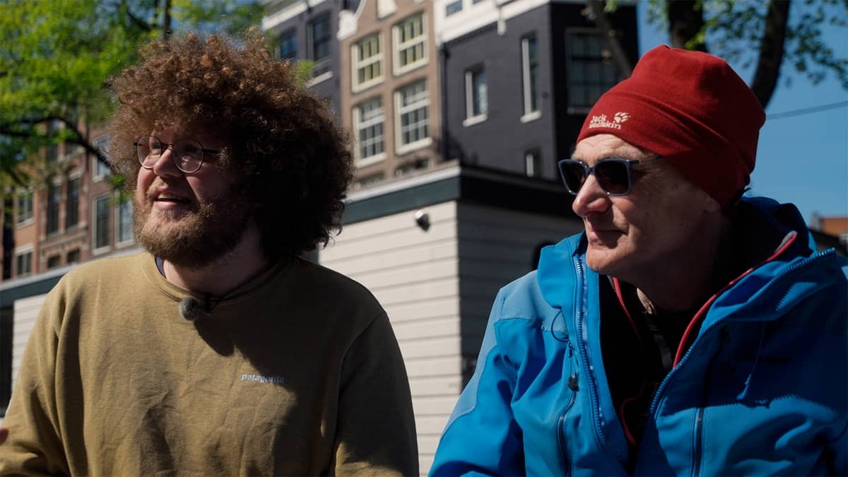 Jonge man met krullen en oude man met muts zitten op een bankje wegkijkend