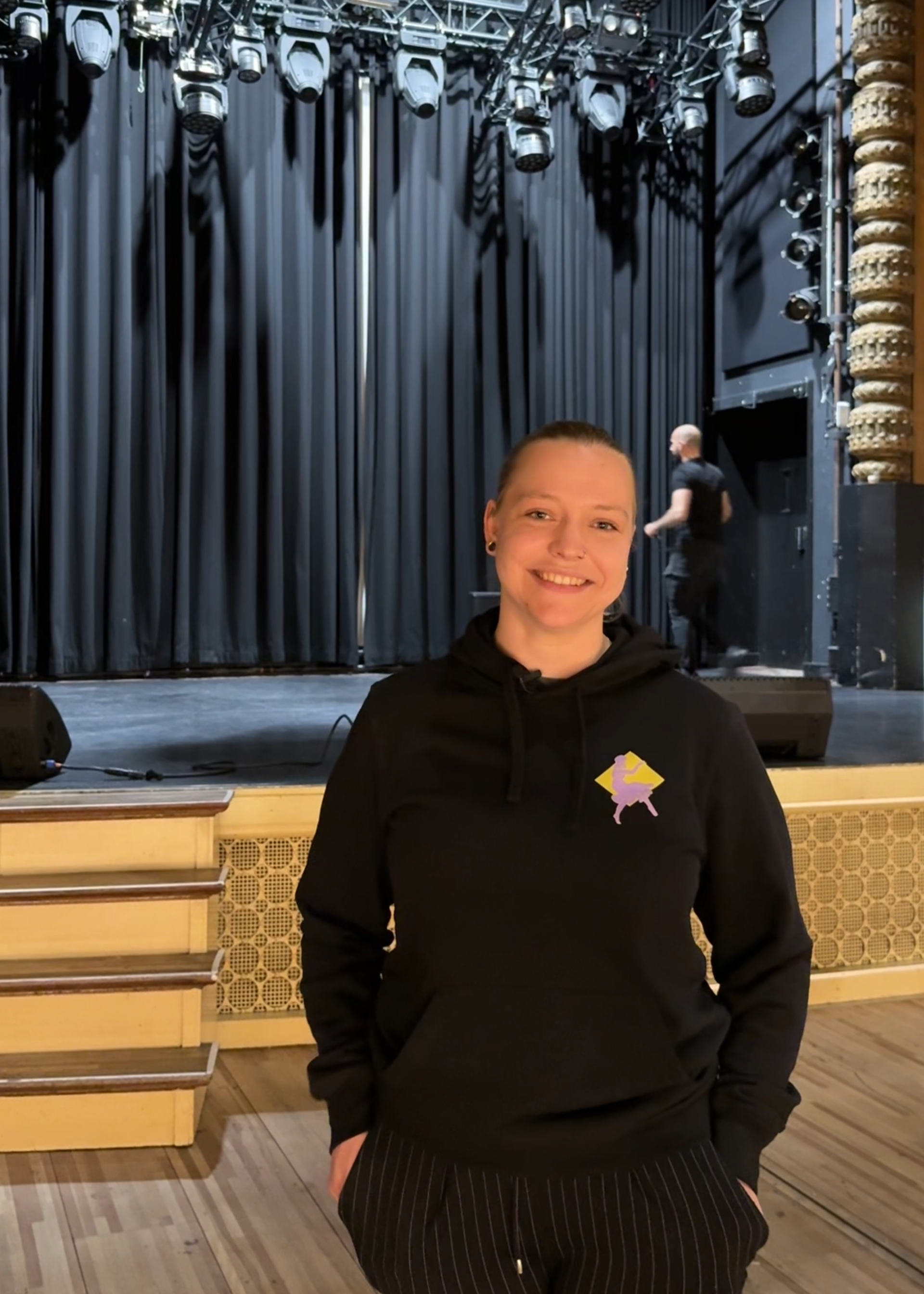 Izanka, de geïnterviewde, in de zaal. Ze heeft zwarte kleren aan en heeft haar handen in haar zakken. Ze kijkt lachend de camera in.