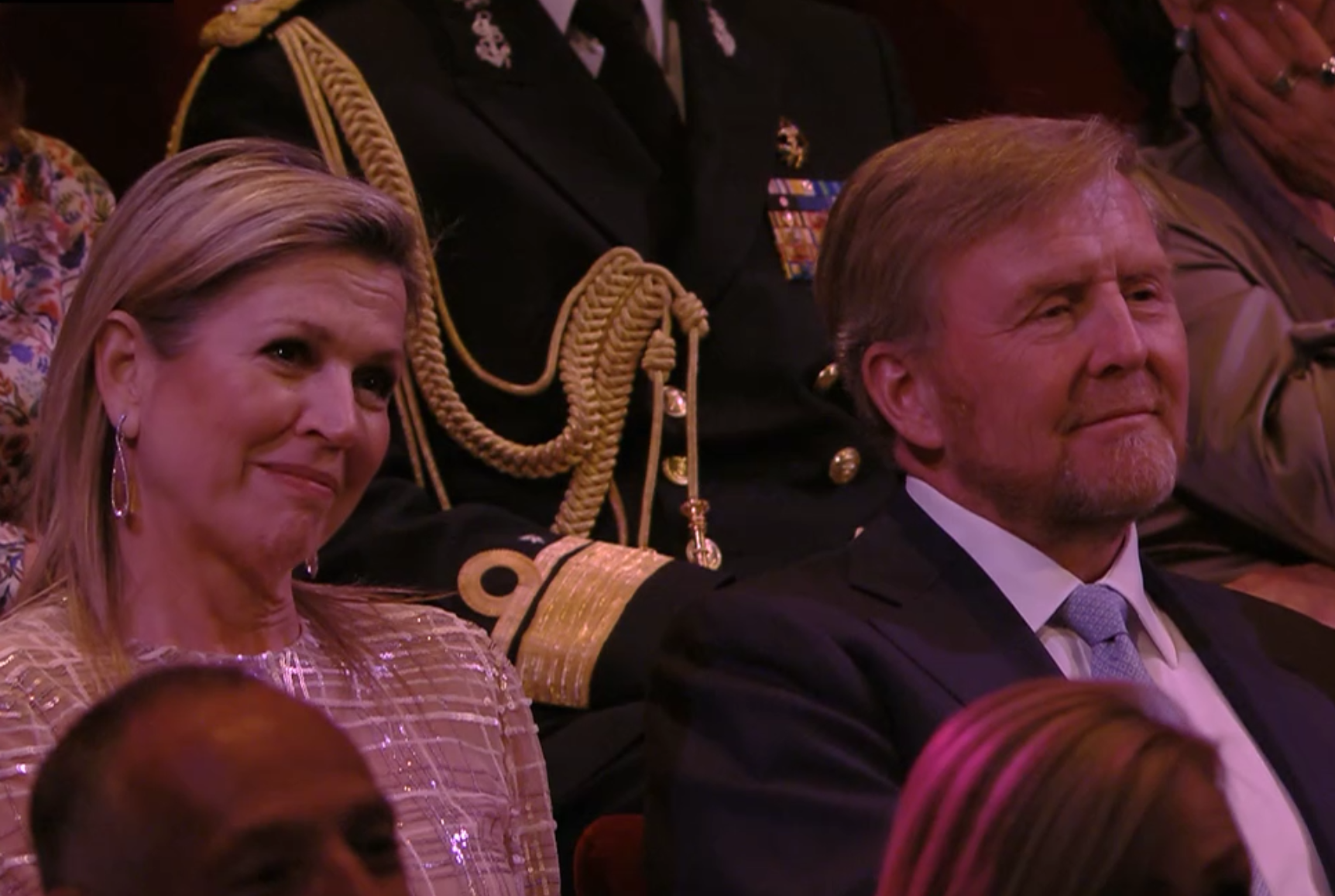 Hier zien we Willem-Alexander en Maxima. Ze zijn netjes aangekleed en zitten in een theaterzaal op rode stoelen en kijken naar een concert buiten beeld.