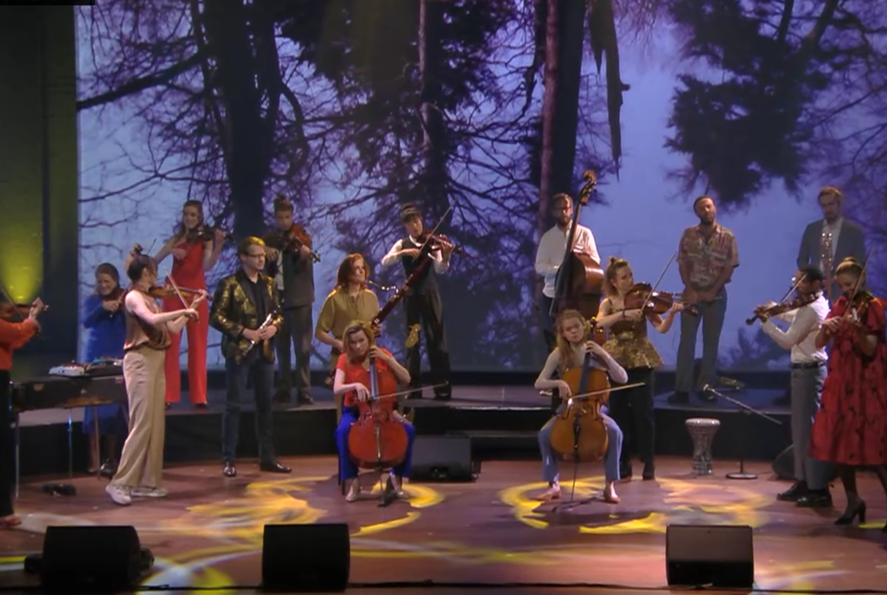 Hier zien we Pynarello op een podium. Het is een groot ensemble met veel strijkers. Vooraan zitten mensen die cello spelen en daaromheen staan mensen met violen in hun handen te spelen.