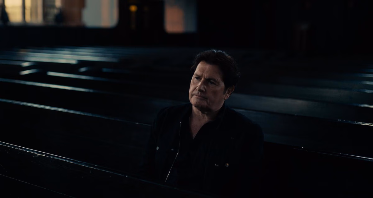Een stil uit de docu, waar zanger Jim Kerr in een theater op een stoel zit. Het is een hele donkere foto. Hij heeft donker haar en kijkt serieus naar iets buiten beeld.