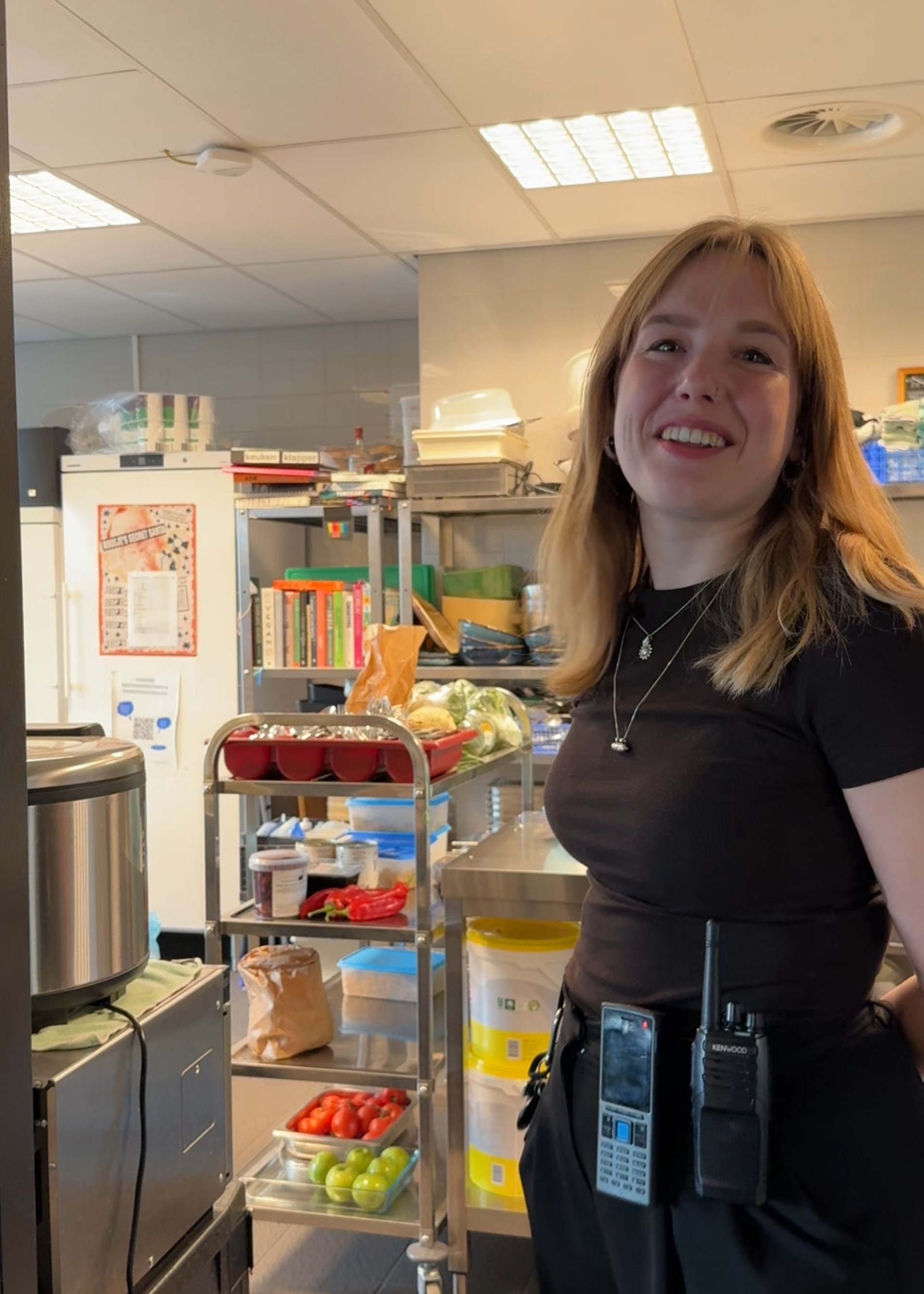 Een foto van Susanne die bij een keuken staat. Susanne is een jonge meid met los blond haar en ze kijkt lachend naar de camera. Achter haar zie je een grote professionele keuken.