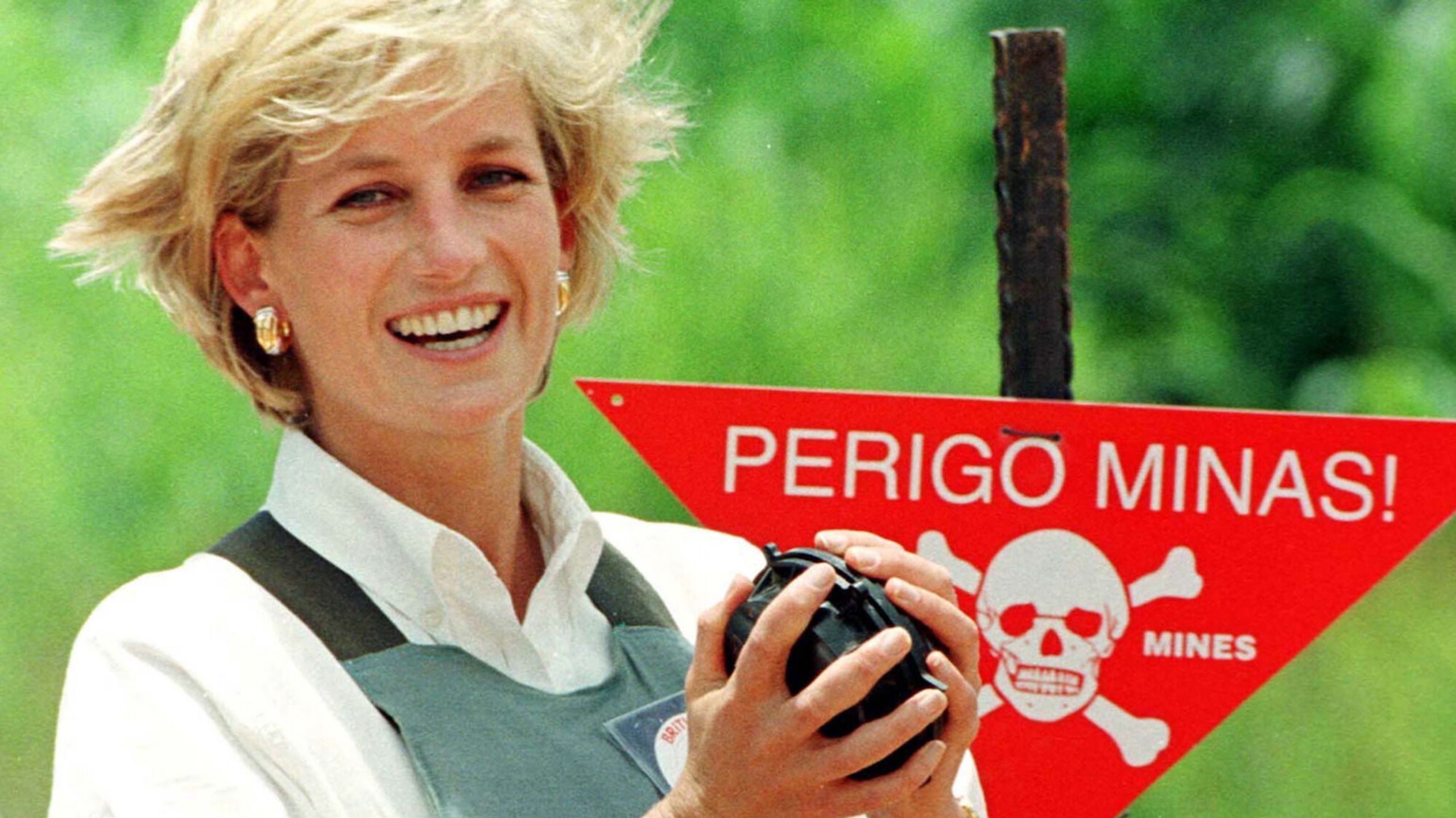 Princess Diana Spencer lacht naar de camera met een granaat in haar handen. Ze staat voor een mijnenveld-bord.