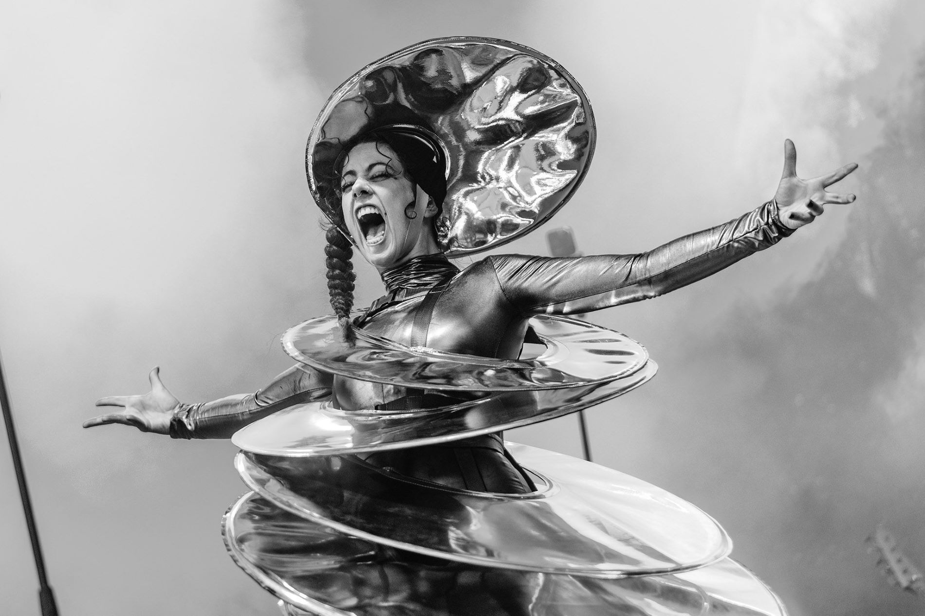Zwartwitfoto van een vrouw die met haar armen wijd op een podium staat. Haar mond heeft ze wijd open en ze heeft een alienachtig kostuum aan dat bestaat uit een soort schotels. Op haar hoofd draagt ze ook een schotel.