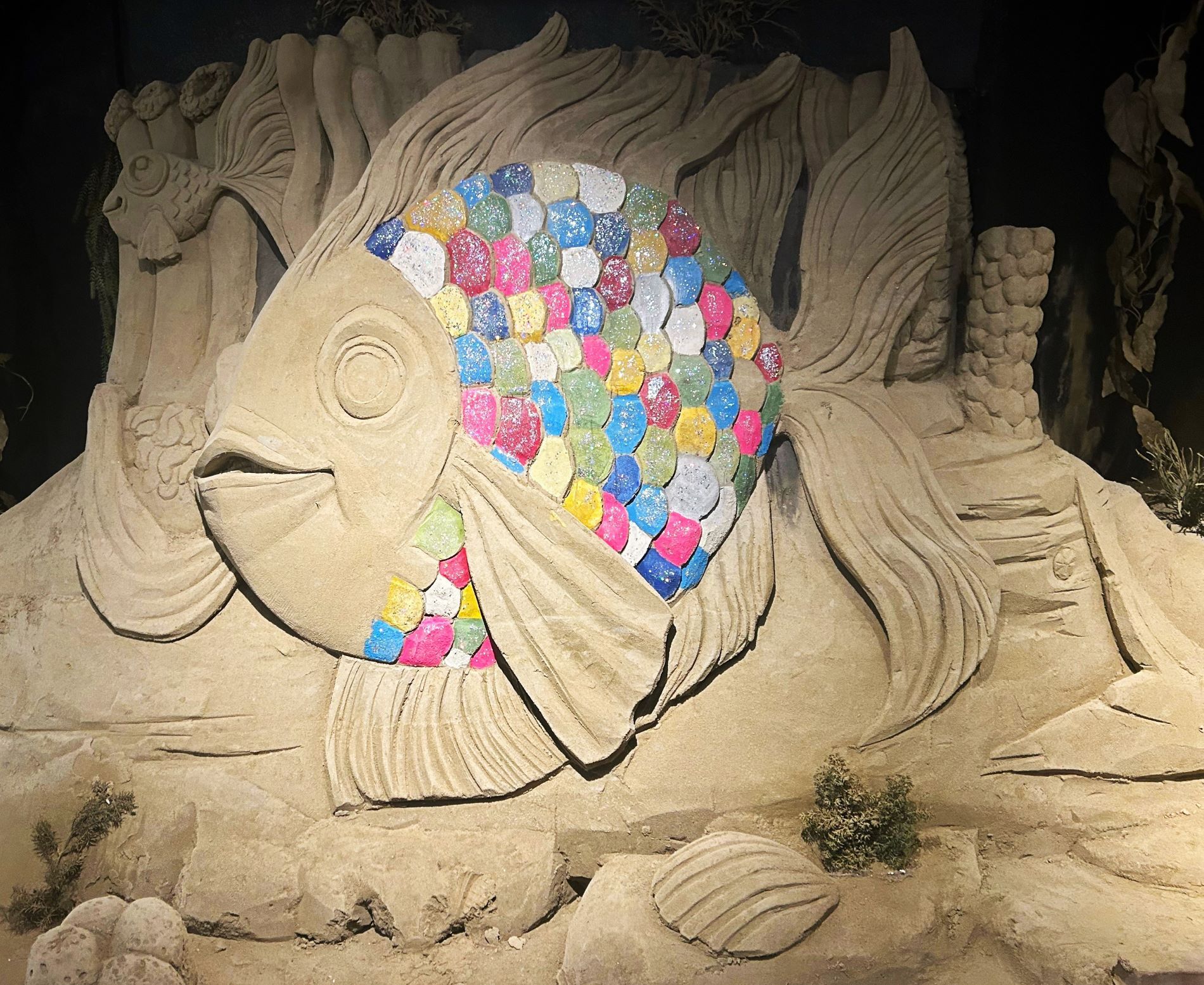 Zandsculptuur van de vis uit het kinderboek 'De mooiste vis van de zee'. Zijn kop en vinnen zijn zandkleurig (beige) en zijn schubben zijn alle kleuren van de regenboog (blauw, roze, groen, rood, wit, geel).